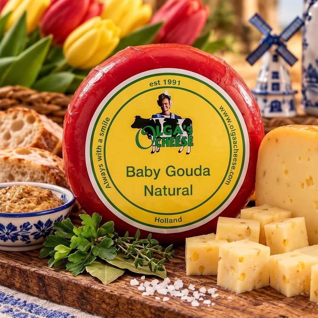 Baby Gouda Natural cheese shown in a round wheel and sliced blocks on a wooden board, with bread, herbs, tulips, and a blue-and-white ceramic windmill jug in a styled kitchen scene. Text on the right describes the cheese as smooth, creamy, and traditional Dutch quality.